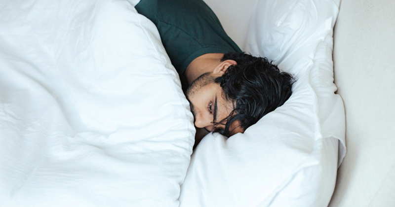 A representation of a man lying in bed under a white comforter with his eyes open
