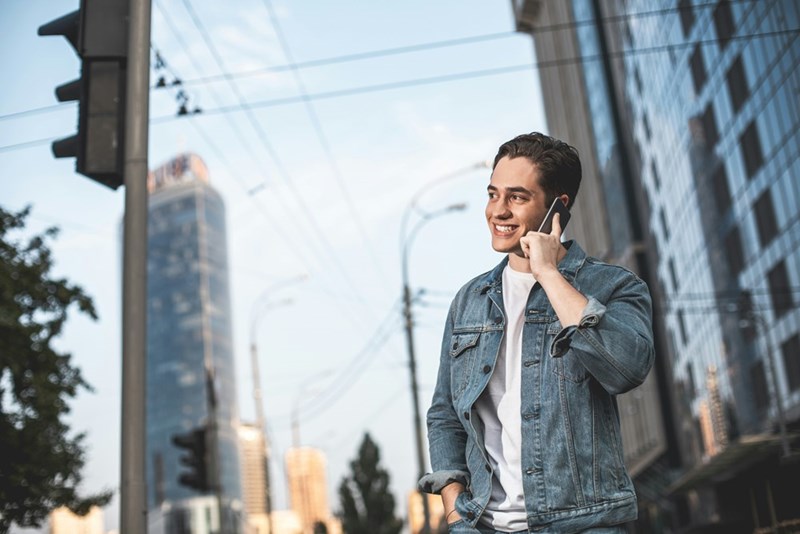 Man in a denim jacket smiles while talking on a phone on a city sidewalk near glass buildings.