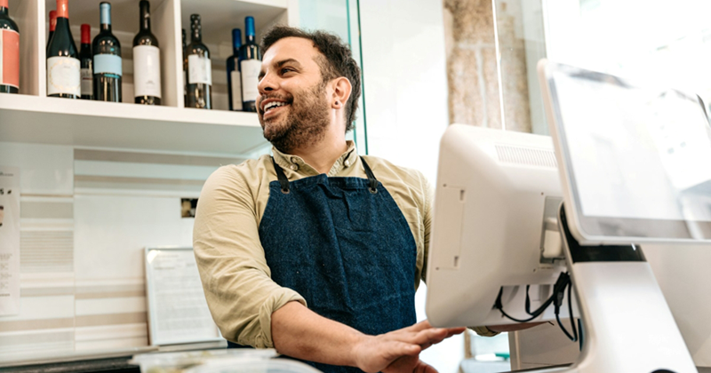 A retail worker looks to the side and smiles while standing behind a touch-screen cash register