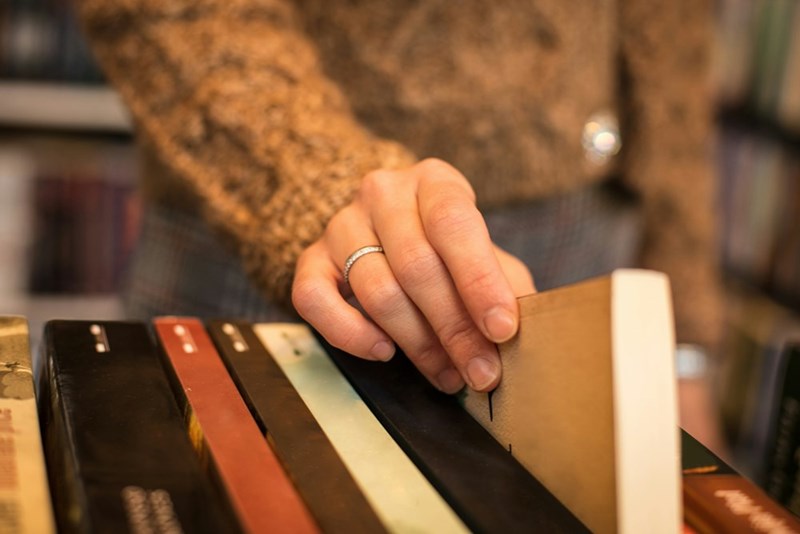 A woman with a wedding engagement ring and a thick sweater picks out a book on a cart at a bookstore.