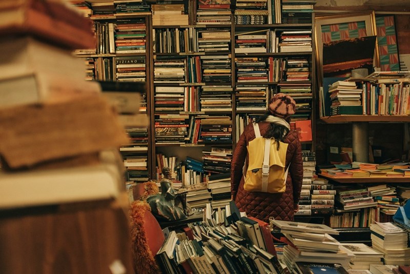 A person with a backpack on and a winter hat looks at a wall of books at a bookstore with piles of books surrounding them.
