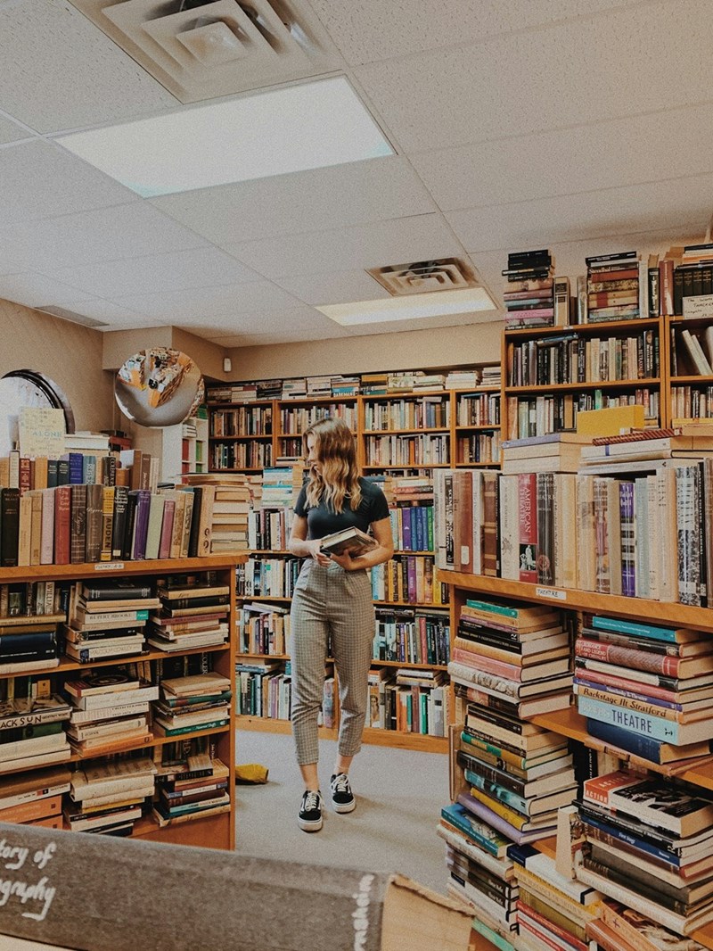 A woman in high-waisted slacks peruses a bookstore with piles of books all around her while she carries a book in her arms.