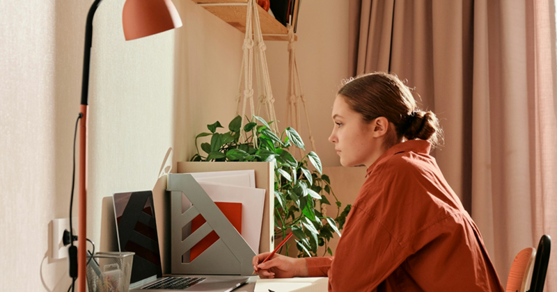 A young woman sits at a table with a laptop, writing in a notepad