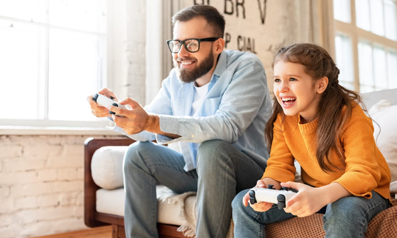 Father and young daughter laughing while playing video games together in the living room.