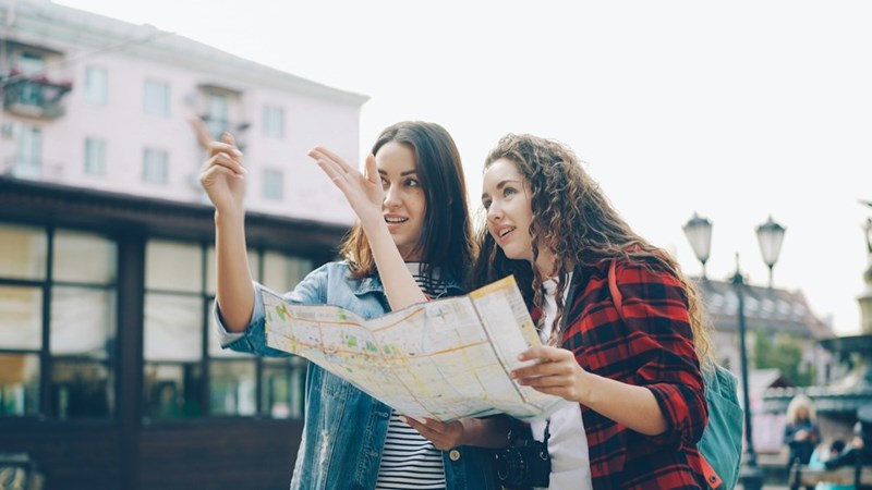 Two women traveling together discuss directions while looking at a paper map in a European city.