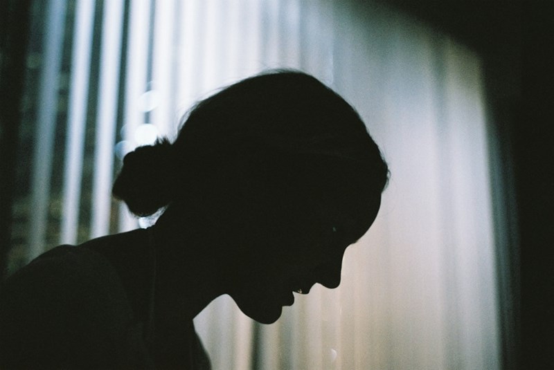 Silhouetted woman sits with her head lowered beside a bright window in a dark room.