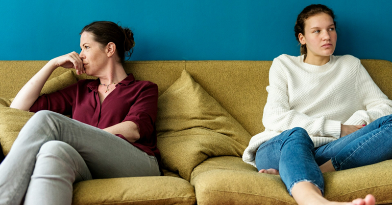 A representation of a mom and her young adult daughter sitting at opposite ends of a couch, looking away from one another