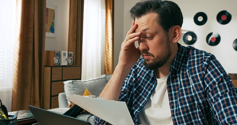 Man in a checkered shirt reviewing papers with his fingers pressed to his forehead, looking weighed down by financial stress