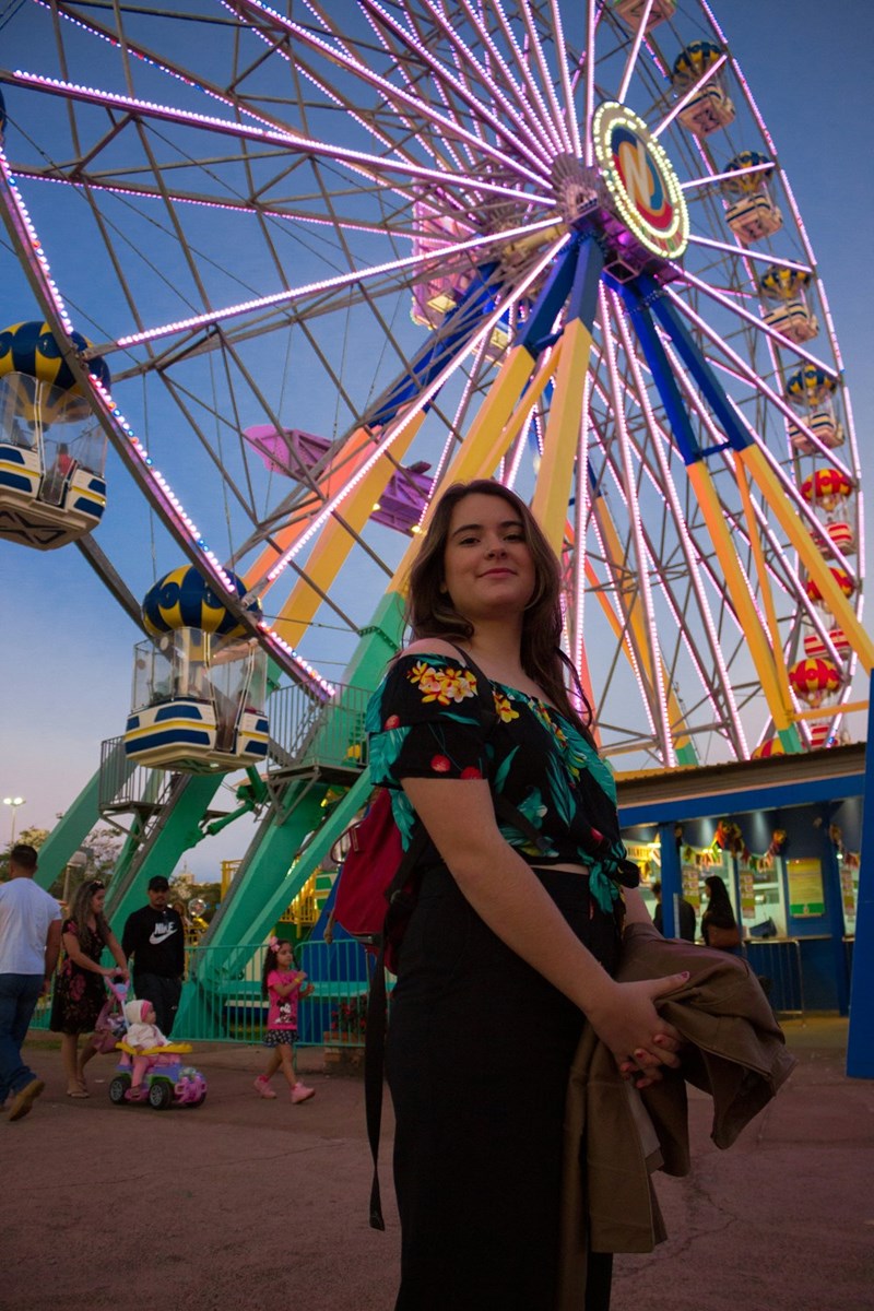 A woman poses smiling in front of a Ferris wheel at an amusement park nearing dusk.