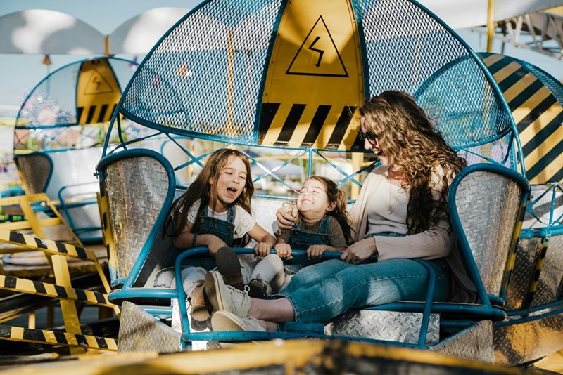 A mom and her two daughters happily laugh and enjoy a spinning ride at an amusement park on a sunny day.