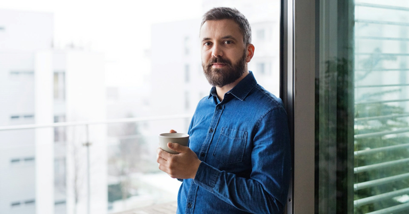 A middle-aged man holding a coffee cup and leaning against a window frame