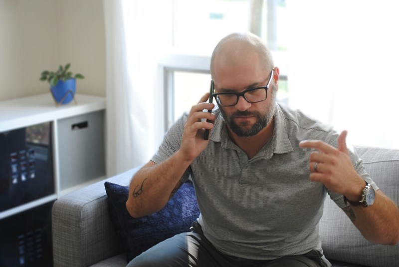 Man talking on a phone while gesturing from a couch, looking serious and frustrated,