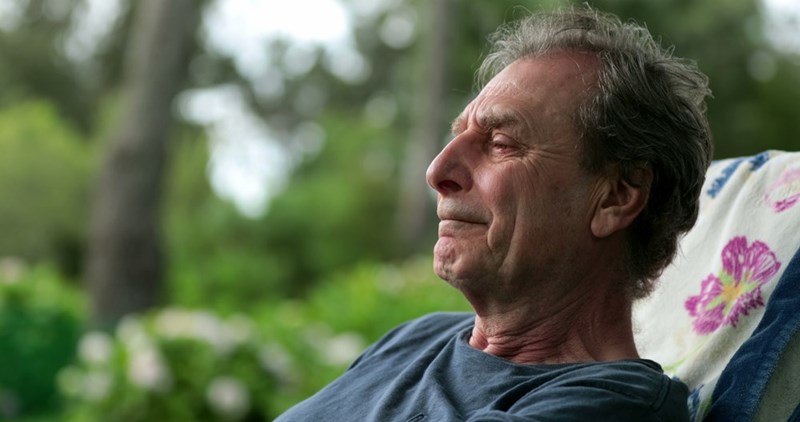 Older man reclines outdoors in a chair, looking pensive with greenery blurred behind him.