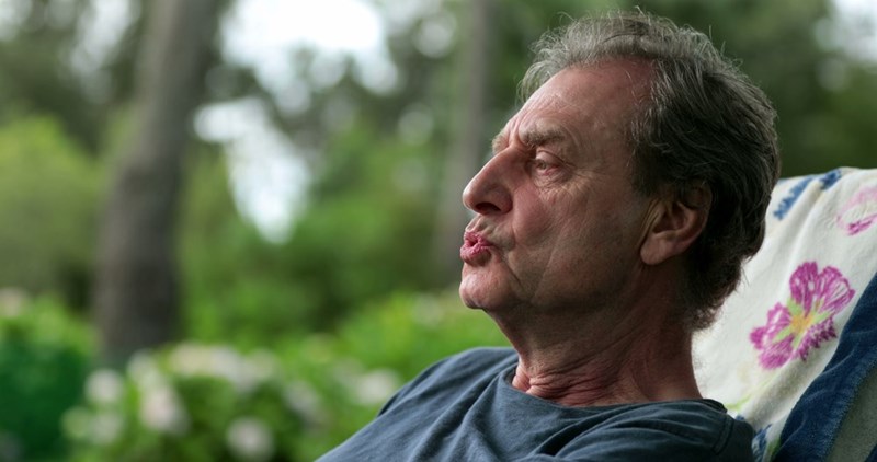 Older man reclines outdoors on a cushioned chair, making a puckered expression with greenery behind him.