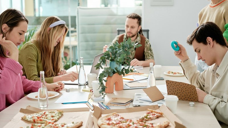Coworkers sit around a meeting table with open pizza boxes, notebooks, water bottles, and coffee cups.