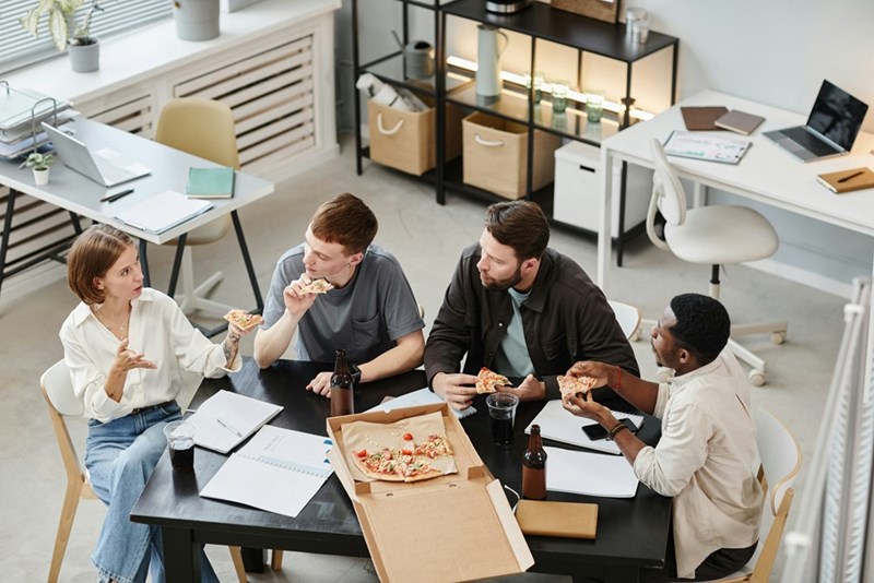 Four coworkers eat pizza around an office table with notebooks, drinks, and an open pizza box.
