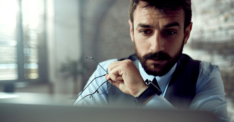 A close-up of a male employee working on a laptop while holding a pair of glasses