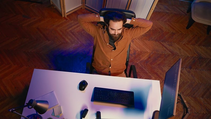 Man leans back with hands behind his head at a desk with monitor, keyboard, mouse, and headphones.