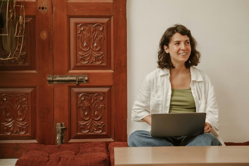 Woman sits on a cushioned bench with a laptop beside a carved wooden door, smiling indoors