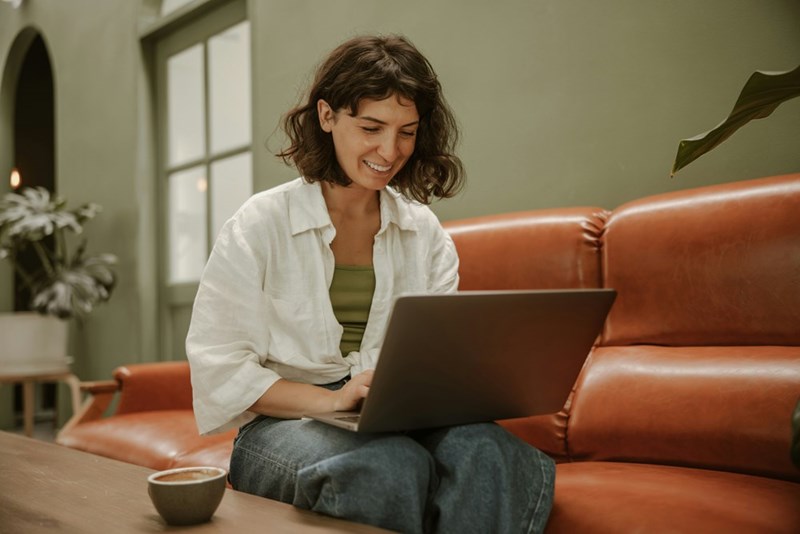 Woman smiles while using a laptop on a brown leather couch with a coffee cup on the table.