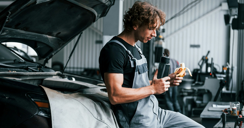 A young male mechanic leans on the open hood of a car, holding a flask and eating a banana