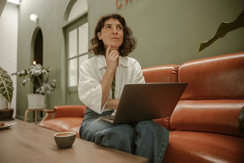 Woman sits on a brown leather couch with a laptop, resting her hand on her chin near a coffee cup.