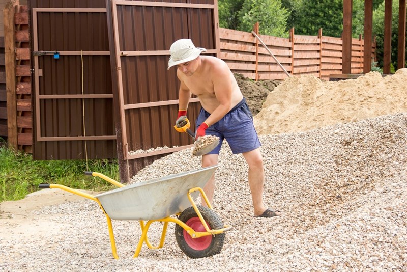 Man shovels gravel into a wheelbarrow near an open gate, fence, and piles of dirt.