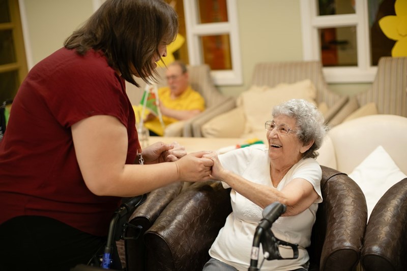 Care home employee warmly greeting and holding hands with an elderly resident in a nursing home lounge, highlighting compassionate senior care, trust, and daily support.