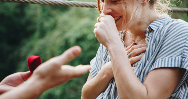 A model representing a woman holding her closed hand to her mouth with her other hand on her chest as she is being proposed to outside
