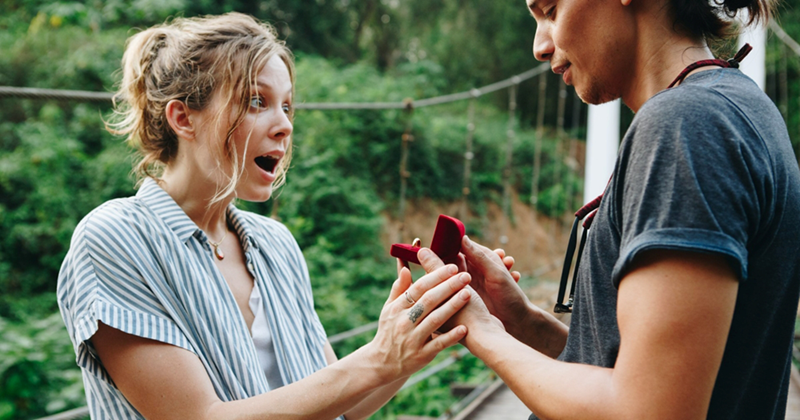 A representation of a woman looking surprised as a man holds out a ring box to her while outside
