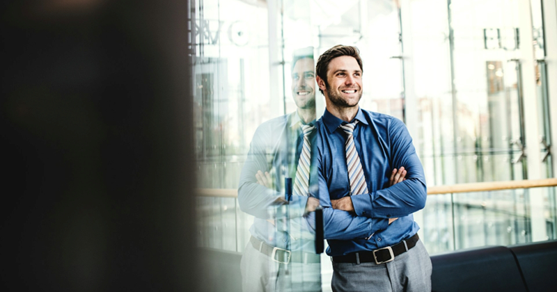 A male business employee smiles and crosses his arms while leaning against the glass wall of an office building