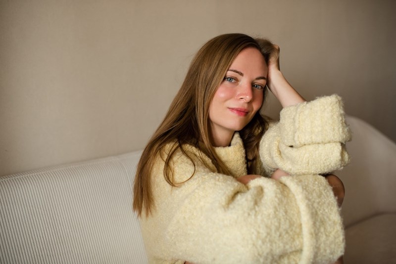 Woman in a cream sweater sits on a light couch with one hand resting in her hair.