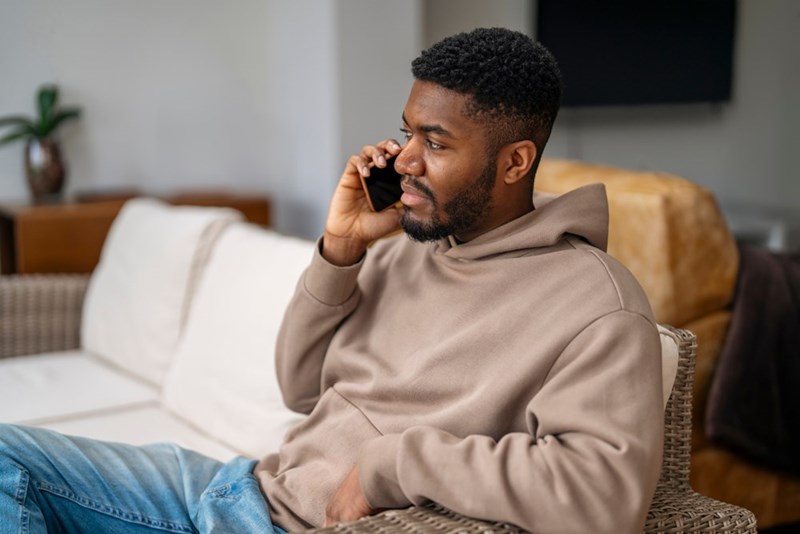 Man in a beige hoodie sits on a couch talking on a phone, looking serious in a living room.