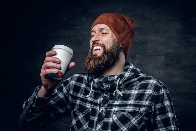 Bearded man in a brown beanie and plaid flannel laughs while holding a takeaway coffee cup.