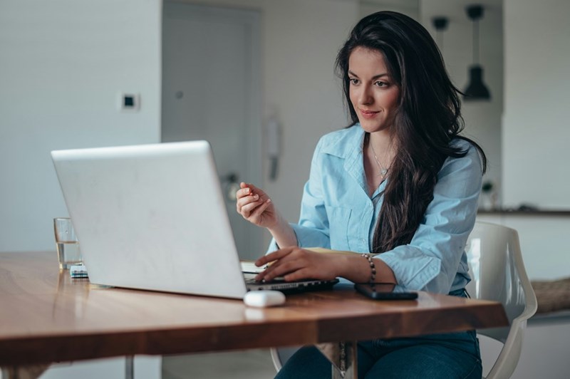 Woman in a blue shirt uses a laptop at a wooden table with a phone, earbuds, and glass of water nearby.