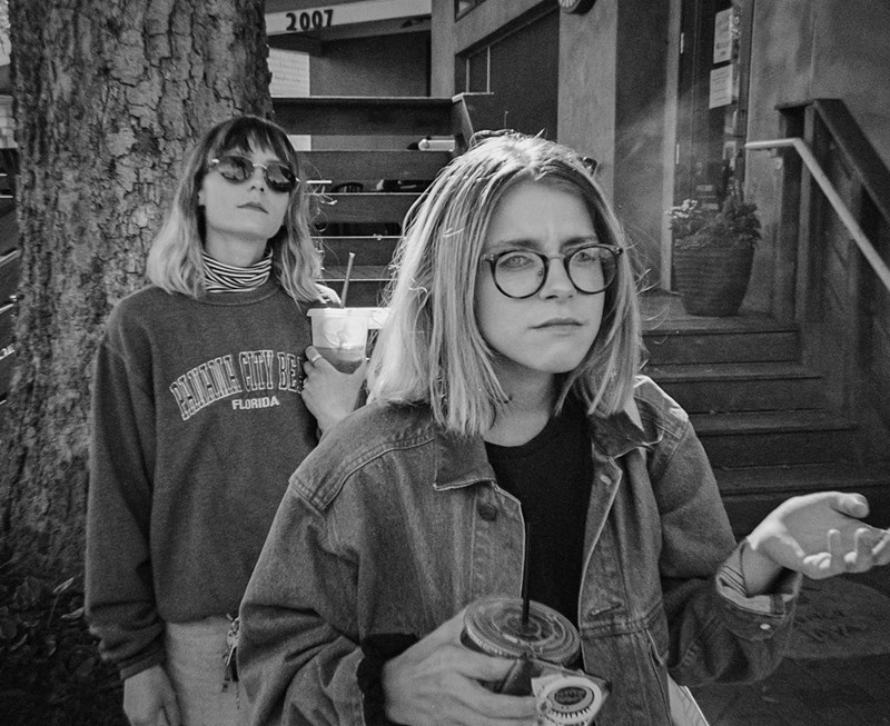 Two young women stand outdoors in black and white, holding drinks