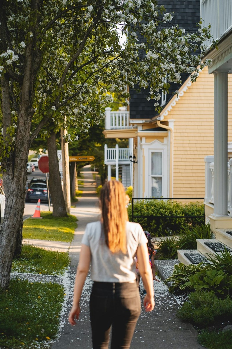 Young woman walks along a residential sidewalk lined with trees, porches, and yellow houses.
