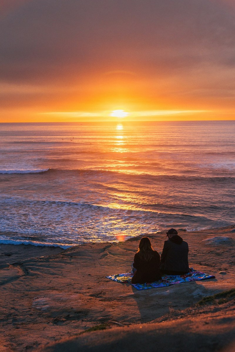 Couple sitting together on a sandy beach at sunset in San Diego, California, illustrating the prepaid birthday trip.