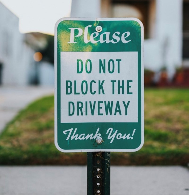 A green and white sign in front of a nice house saying, “Please do not block the driveway! Thank You!”