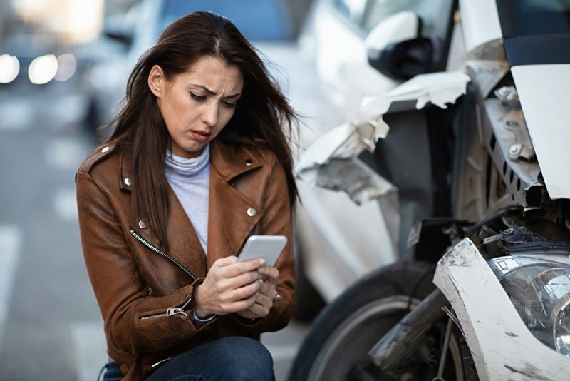Frustrated young woman looking at her phone beside a damaged car.
