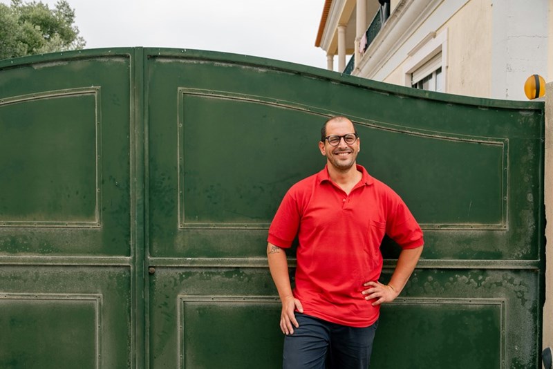 Man in a red shirt stands smiling with one hand on his hip in front of a large green metal gate.