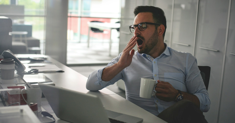 An employee sitting at his work desk yawns while holding a mug and wearing glasses
