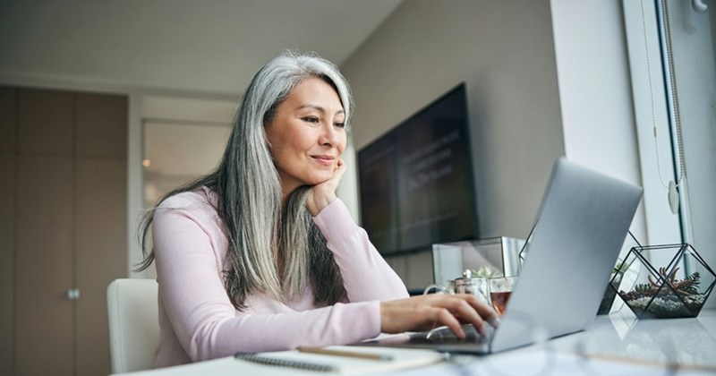 A woman with long grey hair rests her head in her hand as she sits at a table and browses on a laptop