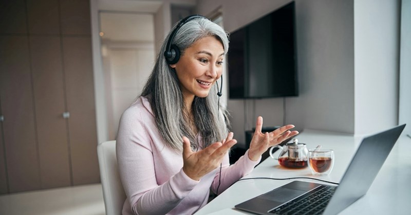 A woman with long grey hair wears a headset and gestures to her laptop with a pot of tea beside her