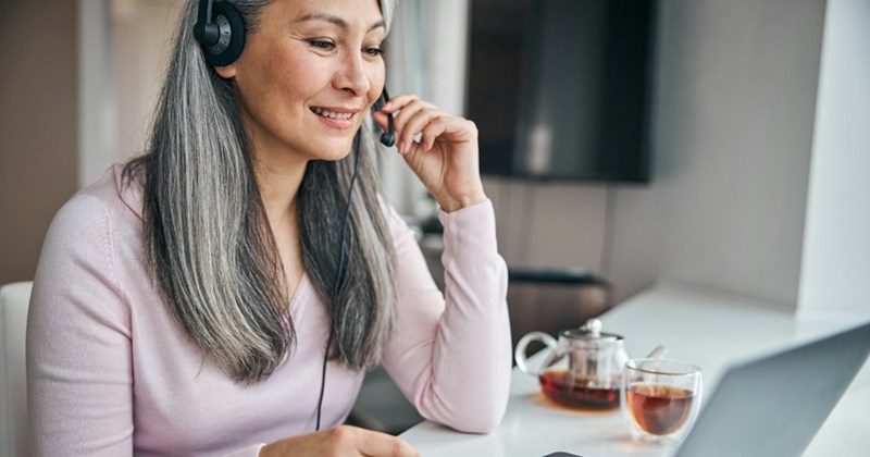 A woman with long grey hair adjusts the mouthpiece on her headset while using a laptop