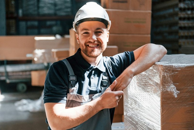 logistics worker smiling in a warehouse while leaning on a wrapped cardboard box.