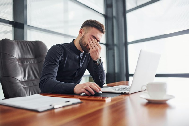 A stressed worker presses his fingers to his eyes at his desk in a bright office
