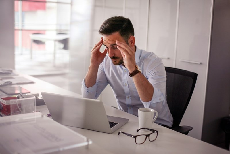 An anxious worker massages his temples at his desk with his glasses on the desk next to him and a coffee mug too