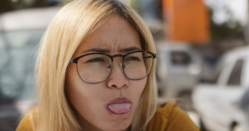 A close-up of a young woman wearing glasses, sitting outside and sticking her tongue out slightly