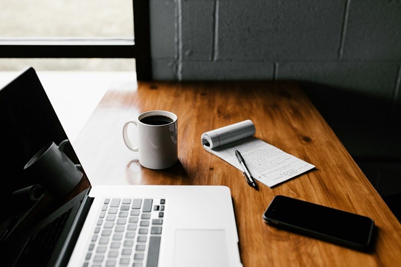 A computer, a cup of coffee, a cellphone, and a notebook displayed at a wooden table.
